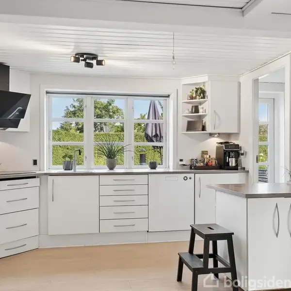 Køkken med hvide skabe og stålgreb, køleskab, ovn og emhætte. Vindue i baggrunden giver udsigt til have. Sort skammel ved siden af en køkken-ø med mørk bordplade.