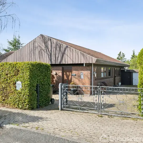 Huset står på en flisebelagt indkørsel, omgivet af velplejede hække og træer. I forgrunden ses en flot smedejernslåge, og bygningen har en rødbrun murstensfacade og rødt tag.