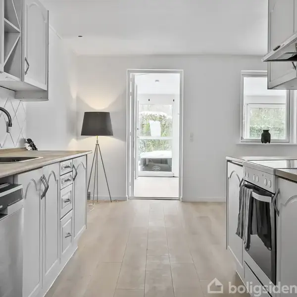 Køkken med hvide skabe og træborde; opvaskemaskine og ovn indbygget; en gulvlampe står tændt; lyse trægulve; en dør åbner til et lyst rum med vinduer og planter.