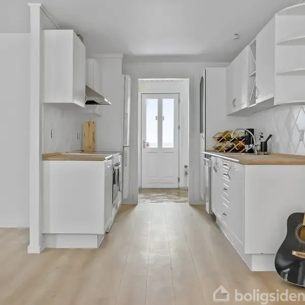 En sort akustisk guitar står op ad et hvidt køkkenbord i et lyst køkken. Køkkenet har træbordplader, hvide skabe og en dør i baggrunden. Ingen tekst til stede.