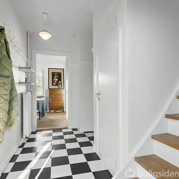 En hvid trappe med trætrin går opad i en entré med sort-hvidt ternet gulv. En grøn jakke hænger på en knagerække, og et værelse ses i baggrunden.