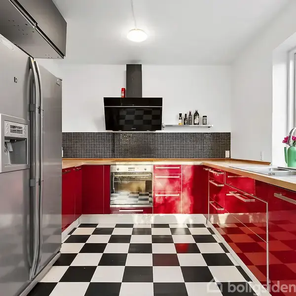 Et moderne køkken med røde skabe og skakternet sort-hvidt gulv. Køleskab og ovn til venstre, vask ved vinduet til højre. Neutral vægfarve med dagslys fra vinduet.