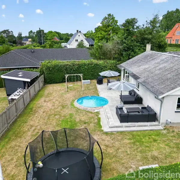 Hus med grå tag, terrasse med møbler og parasol. I haven ses en trampolin, gyngestativ og en rund pool. Omgivet af græsplæne og hegn, med nabohuse i baggrunden.