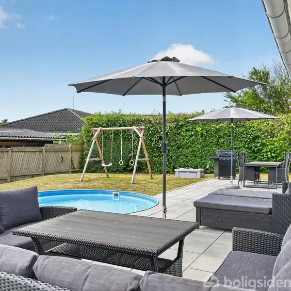 Terrasse med grå sofaer og parasoller, omkranset af et hegn. En rund pool og en gynge ses i baghaven, omgivet af hække. Klar himmel i baggrunden.