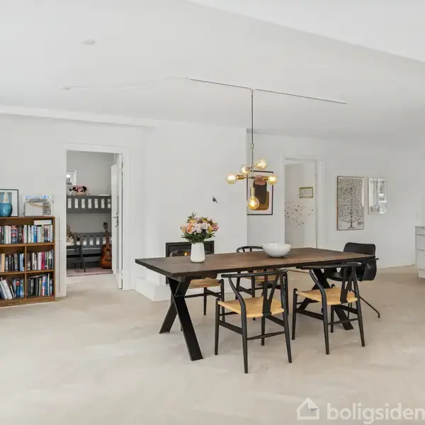 Et moderne spisestueområde med et mørkt træbord og fire sorte stole i et lyst rum. Til venstre står en bogreol ved siden af planter, og til højre ses et køkkenområde med hvide skabe.