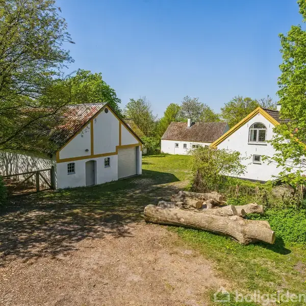 To huse med karakteristiske hvide vægge og gule kanter står i en grøn, skovklædt have. Foran dem ligger en stabel træstammer på en grusbelagt vej.