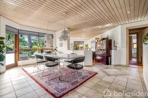 Spisebord med sorte stole står på et tæppe i en spisestue. Store vinduer slipper naturligt lys ind. Rummet har flisegulv og træloft.