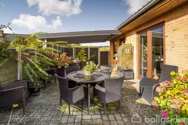 Terrassemøbler placeret rundt om et rundt bord, omgivet af planter og blomster i en solrig have, med udsigt til en murstensbygning og åben dør.