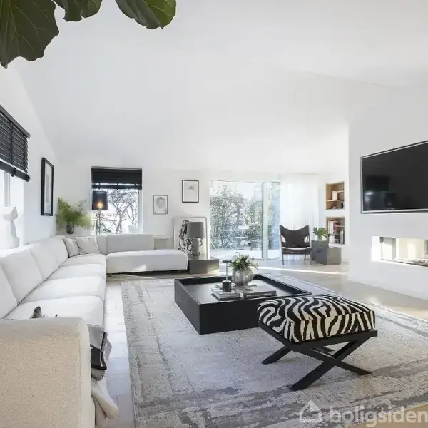 Stue med hvid sofa og firkantet sort bord, stående på lys tæppe. Stort vægmonteret TV, vinduer med sorte rullegardiner. Moderne dekorationer og lys fra terrassedøre i baggrunden.