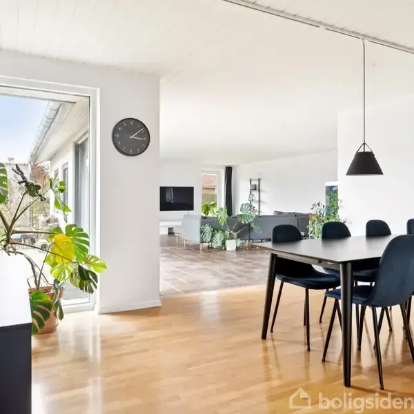 Spisebord med sorte stole står i et lyst, minimalistisk rum med trægulv. Tre hængelamper er placeret over bordet. En væg har ur, planter og stort vindue.