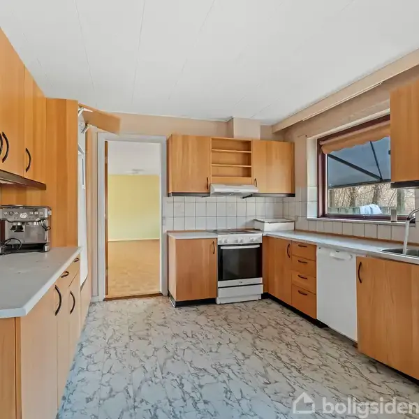 Køkken med brune skabe og hvide fliser, har en kaffemaskine, komfur, opvaskemaskine, vask og vindue med udsigt til træer. Lysegråt, mønstret gulv og dør til tilstødende rum.