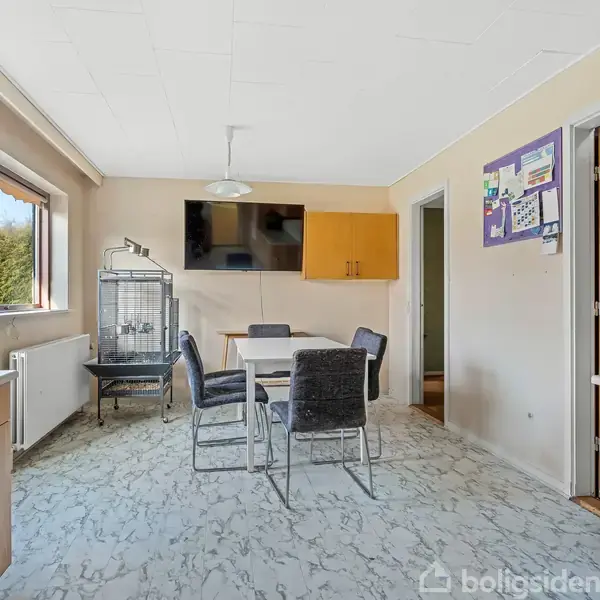 Et køkken med et bord og fire stole i midten, en fuglebur ved siden af vinduet, vægskab og opslagstavle i baggrunden. Ingen tekst findes i billedet.