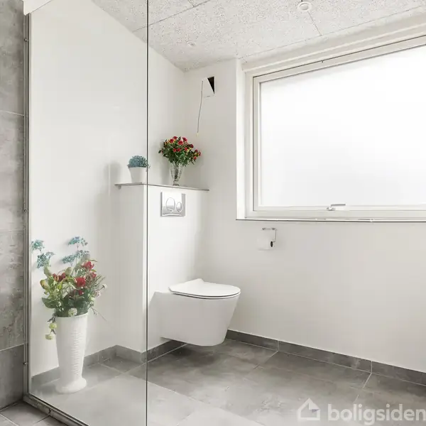 Toilet hænger på væggen under et vindue i et lyst badeværelse. Hylde med blomster over toilettet, og en vase med blomster står på gulvet. Bruser synlig til venstre.