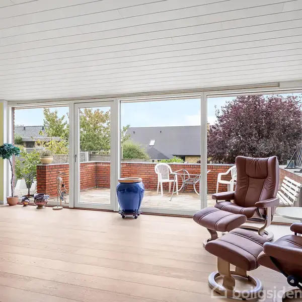 To komfortable lænestole med skammel står i en lys stue med trægulv, vendt mod store vinduer og en terrasse. Udenfor er der en blå krukke og et træbord med stole.