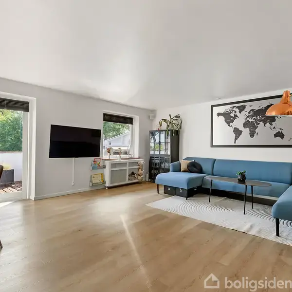 Stue med blå sofa, hvor en verdenskortplakat hænger ovenover. Et kobbergulvlampe står ved siden af. Fjernsynet er monteret på væggen, der er store vinduer mod en altan.