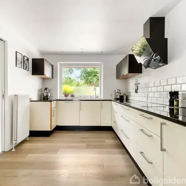 Et moderne køkken med hvide skabe og sort bordplade. Rummelig arbejdsflade, indbygget ovn, og udsigt til en grøn have gennem et vindue. Lyse trægulve og minimalistisk indretning.