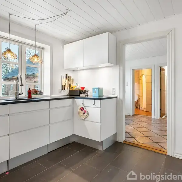 Et lyst, moderne køkken er indrettet med hvide skabe og sorte bordplader. En stor vindue lader dagslys ind, og hængelamper kaster lys over vasken. En åben dør fører til et flisegulv.