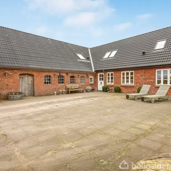 Gårdhave med brostensbelægning, flankeret af røde murstensbygninger med skrå sorte tage. Indrettet med bænke og liggestole. Blomsterkrukker og trædør tilføjer karakter til den åbne gårdplads.