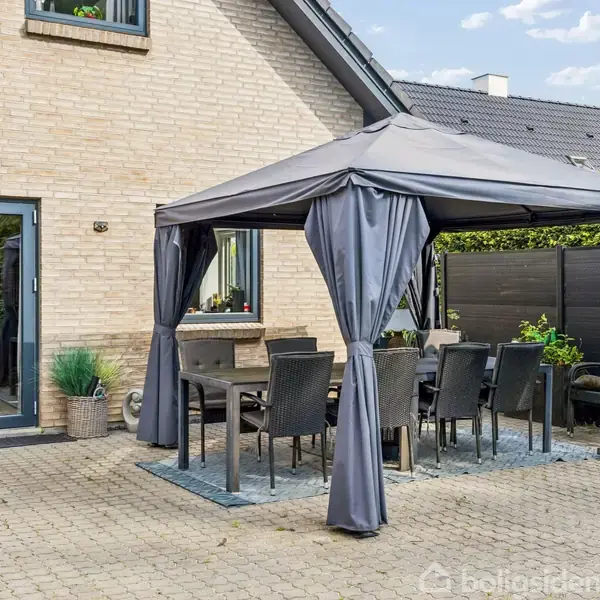 Et gråt udendørs pavillon-telt dækker et bord med seks sorte stole. Pavillonen står på en terrasse af lyse grå fliser ved siden af et murstenshus med grå vinduesrammer.
