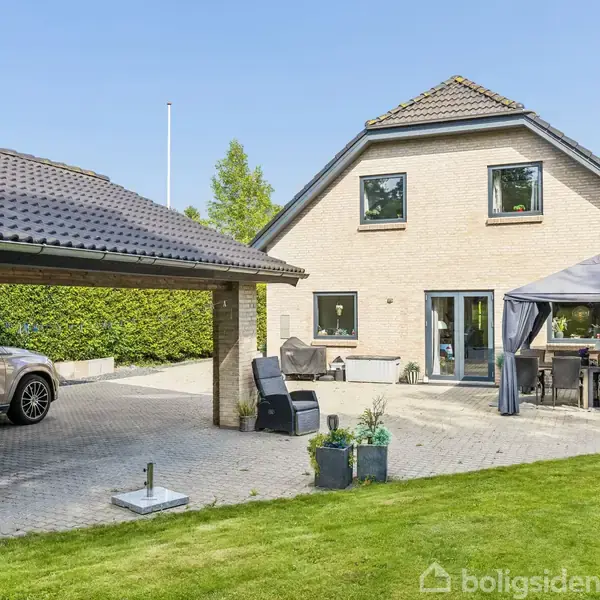 Et to-etagers hus med grå tagsten står i en have. Foran ses en terrasse med et havebord under en pavillon. Til venstre holder en bil under en carport.