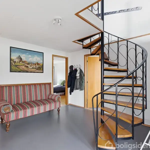 En snoet trappe i metal og træ fører opad. En stribet sofa står til venstre, omgivet af lyse vægge, døråbninger og et billede på væggen.