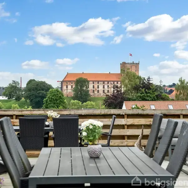 Udendørs terrasse med sorte havemøbler og blomster på et bord. I baggrunden ses et slot og et kirketårn omgivet af grøn beplantning under en blå himmel med spredte skyer.