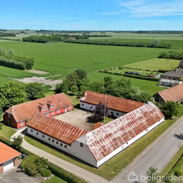 Gårde omgivet af marker ses ovenfra. Røde tagsten og hvide vægge dominerer bygningerne, som danner en firkantet gårdsplads. Området er landligt med åbne marker og få træer.