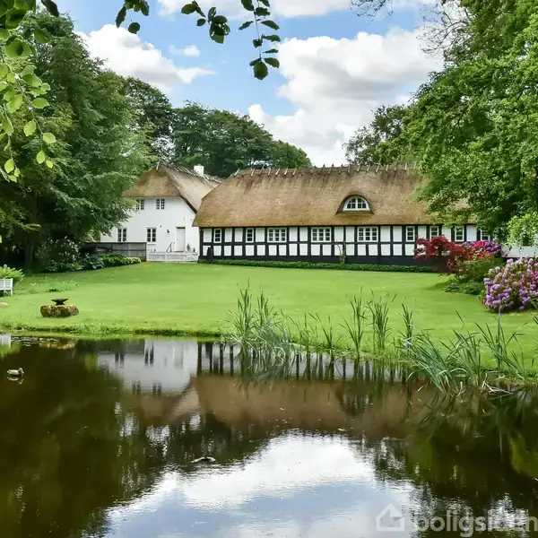 Stråtækt bindingsværkshus står stille foran en velplejet græsplæne og farverige blomster. Området er omgivet af frodige træer og en rolig dam i forgrunden.
