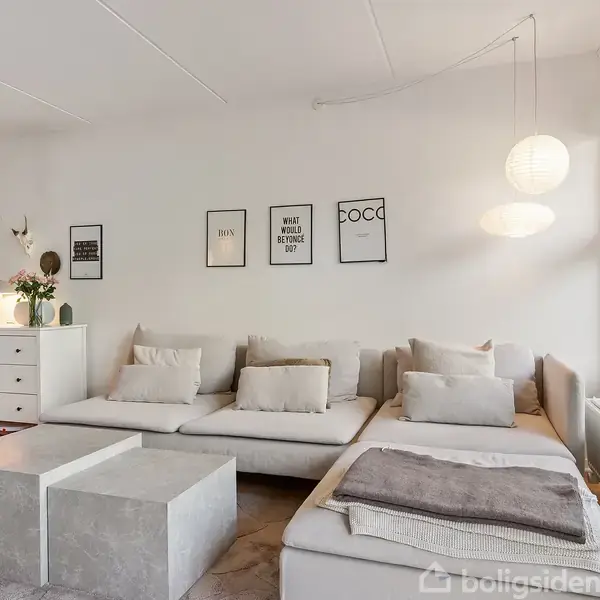 Stue med grå sofa og to firkantede borde. På væggen hænger billeder med tekst: "HUS," "What we do in life echoes in eternity," og "COCO." To lamper hænger fra loftet; store vinduer til højre.
