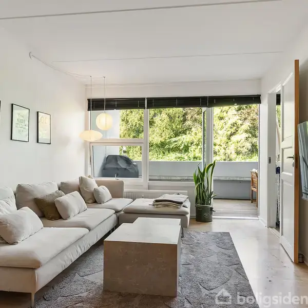 En beige sofa står i en stue ved siden af et sofabord; billeder hænger på væggen over sofaen. Store vinduer viser grøn vegetation udenfor. En fladskærm hænger på væggen.