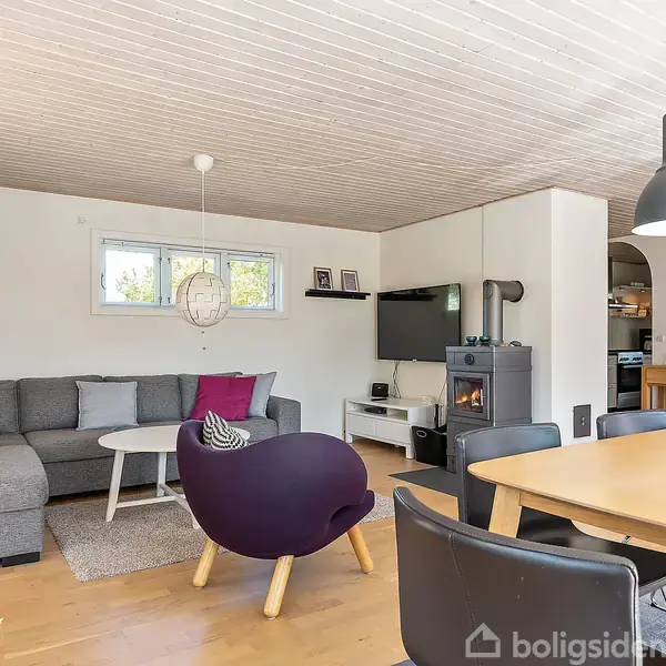 Grå sofa med farverige puder står i hjørnet foran et lille bord. Ved siden af er en mørk lænestol. Rummet har store vinduer, et spisebord og en brændeovn.
