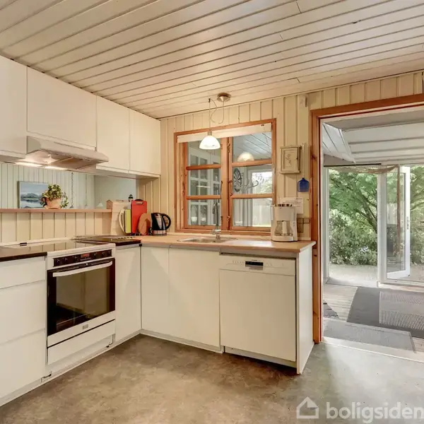 Et lyst køkken med hvide skabe, hvor en ovn og opvaskemaskine er indbygget. Et vindue og en dør oplyser rummet, fører ud til en overdækket terrasse med grønt udsyn.