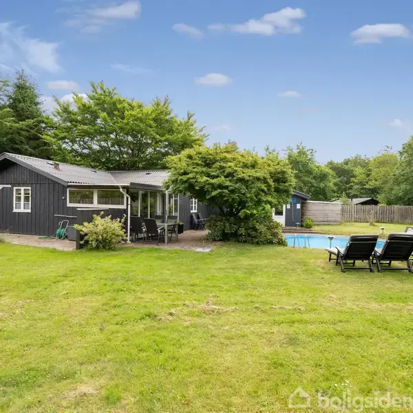 Et sort træhus med veranda står ved en pool. Omkring er der grønne træer og buske. Flere liggestole er placeret på en græsplæne i en fredelig have.