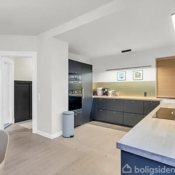 Et moderne køkken med mørke skabe og integrerede apparater. En bordplade med vask er til højre. Billeder hænger over skabene, lys skinner fra en loftslampe.