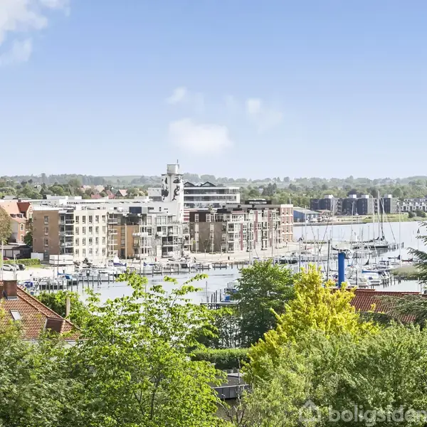 Havn med fortøjede både ligger ved siden af moderne bygninger. Omkring havnen ses grønne træer og buske, mens der i baggrunden er en skovdækket bakke og en klar himmel.
