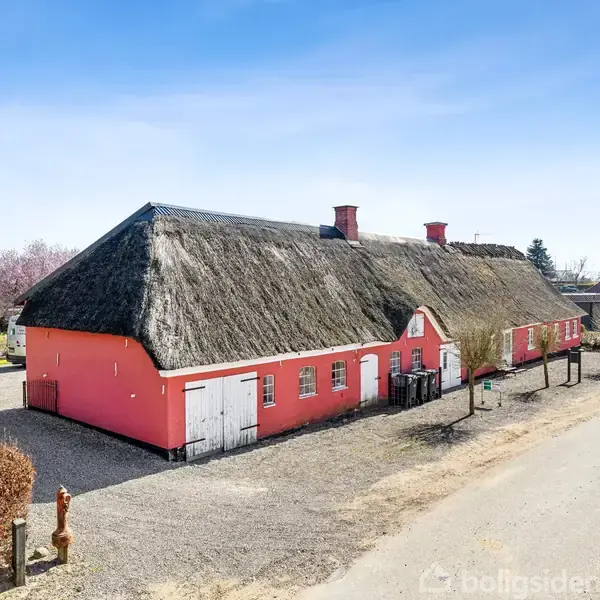 Rødt hus med stråtag og hvide døre står stille på en grusvej. Omgivet af græsarealer og et andet hus i baggrunden, under en klar blå himmel.