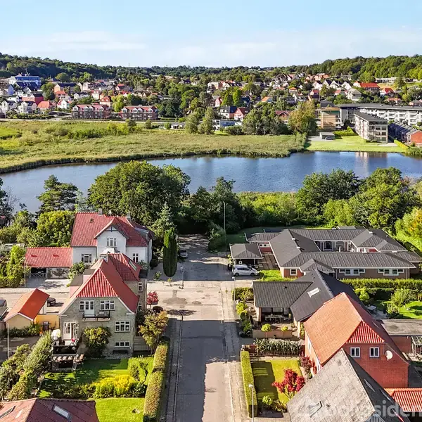 Røde tage på huse grænser op til en rolig sø omgivet af grønne træer, med en by i baggrunden. Klar himmel og frodig natur skaber en idyllisk landskab.