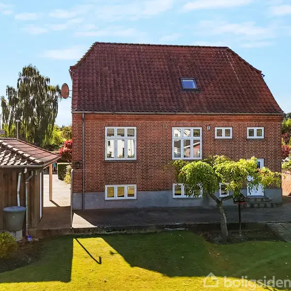 Rødt murstenshus med symmetriske vinduer står stille i en solrig have; træer og buske omringer huset, der har en rød tegltag og ligger mellem andre villaer.