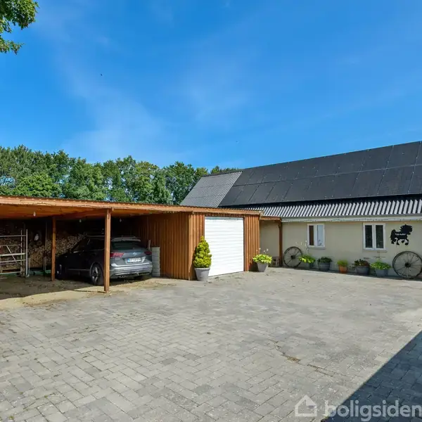 En bil holder parkeret under en overdækket garage ved siden af en hvid bygning med sort tag. Omkring bygningen er der grønne træer og en flisebelagt gårdsplads under en klar blå himmel.