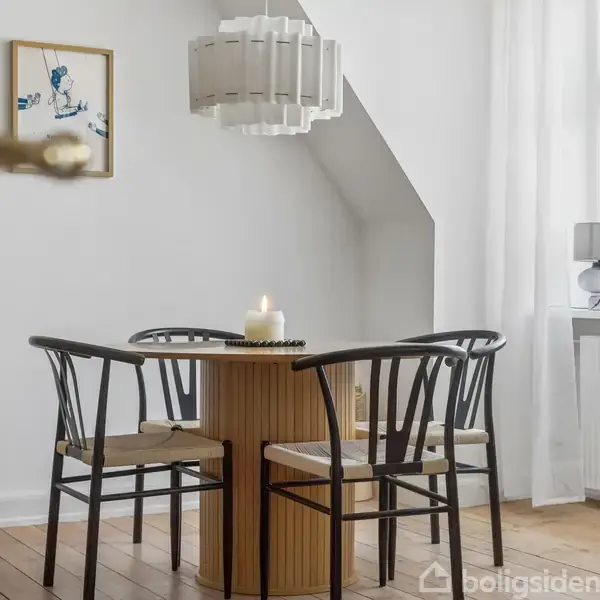 Et rundt spisebord med fire sorte stole står på trægulvet. En tændt stearinlys er midt på bordet. Rummet har hvidmalede vægge, et vindue med hvid gardin og et maleri.