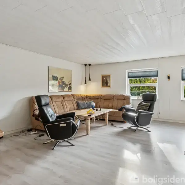 Stue med en brun hjørnesofa, to sorte lænestole og et trækkaffebord. Store vinduer giver lys, og væggene er dekoreret med malerier. Lyst gulv og lyst møblement.