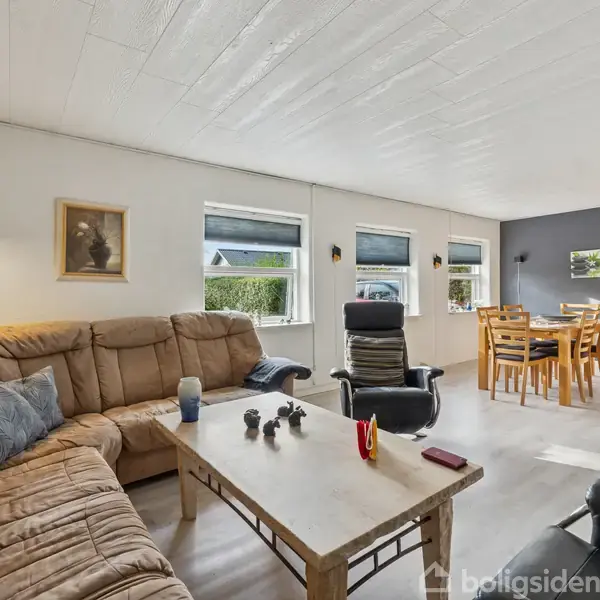 Brun sofagruppe står ved et rektangulært sofabord, dekoreret med små figurer og lys, i en lys stue. Spisebord med stole er i hjørnet ved vinduer.