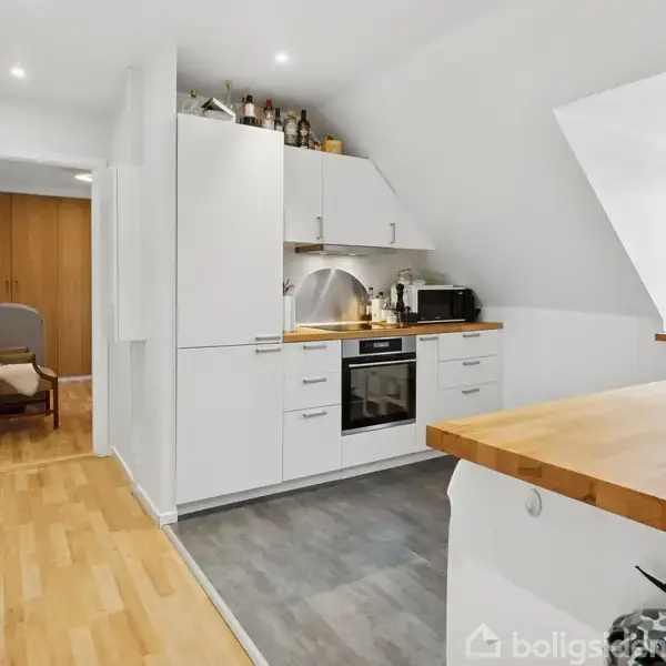 Et moderne køkken med hvide skabe står i centrum. Ovn og mikroovn er integreret. Bøger og flasker står på toppen af skabene. Rummet er lyst med trægulv og lille spiseplads.