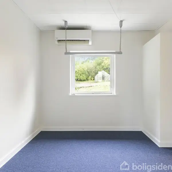 Rummet indeholder en enkelt lampe og et klimaanlæg over et vindue, der giver udsigt til en grøn have med buske og træer. Væggene er hvide, og gulvet er dækket af blåt tæppe.