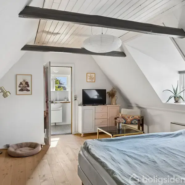 Soveværelse med seng i forgrunden og skråt loft med synlige bjælker. Et møbel med TV står foran, ved siden af en åben dør til badeværelse. Plante i vindueskarmen.