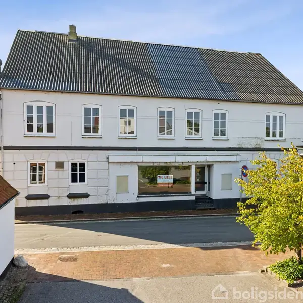 En hvid to-etagers bygning med sort tag har flere vinduer, et butiksvindue med skiltet "TIL LEJE", og står ved en vej. En grøn, frodig busk ses til højre.
