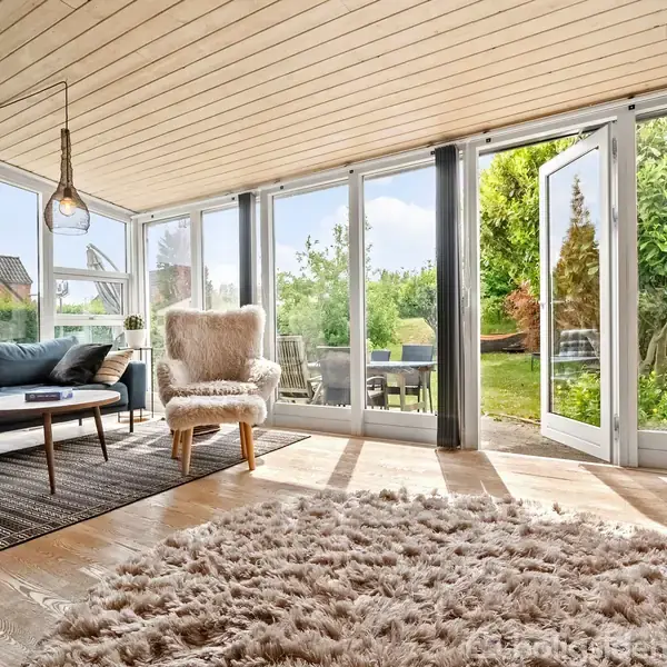 En stue med en mørk sofa og en pelset lænestol står på et trægulv med et tæppe. Store glasdøre åbner ud til en frodig have.