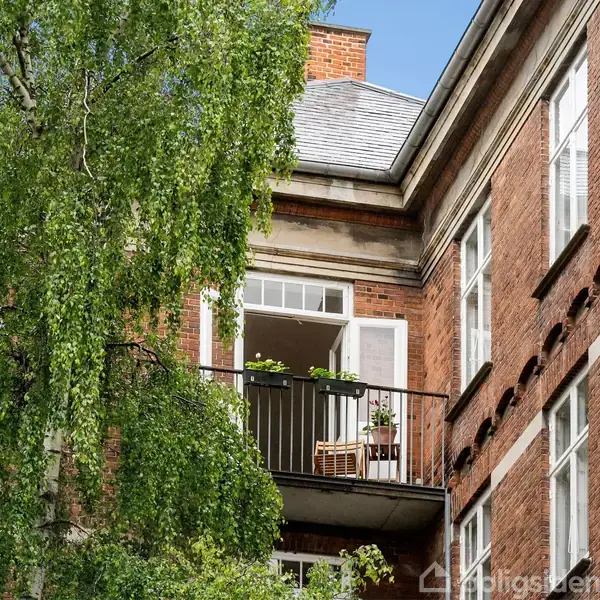 En balkon med en åben dør og et grønt træ foran, omgivet af en rød murstensbygning med flere vinduer i et urbant miljø.