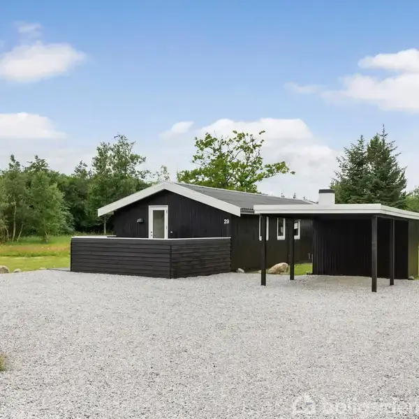 Sort sommerhus står stille på en grusplads omgivet af grønt græs og træer. Det har en terrasse og et lille halvtag. Himlen er blå med lette skyer.