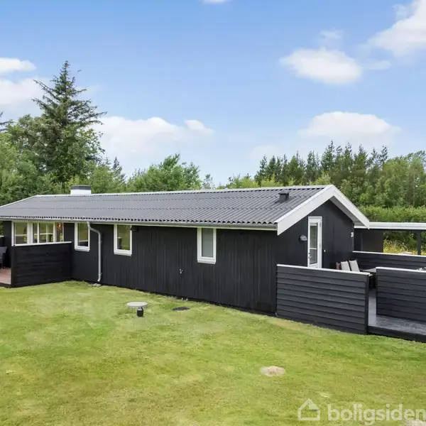 Sort træhus med terrasse står stille på en grøn græsplæne, omgivet af træer under en blå himmel med spredte skyer.
